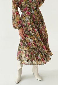 Frau in einem langen Blumenkleid mit rosa, gelben und weißen Blumen, kombiniert mit weißen spitz zulaufenden High-Heels.