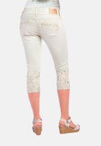 Weiße Caprihosen mit floralem Stickerei an den Säumen, kombiniert mit Wedgesandalen mit einem gemusterten Riemen. Denim-Material in taillierter Passform.