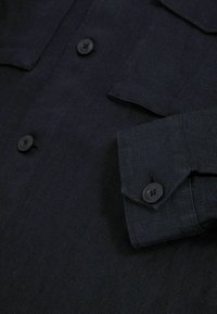 Cămașă button-up de culoare bleumarin închis, realizată dintr-un material texturat. Are două buzunare pe piept și închideri cu nasturi negri.