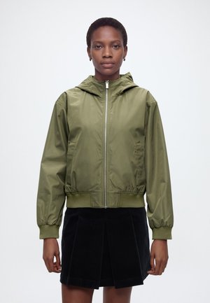 Frau trägt eine olivgrüne Kapuzenjacke mit Reißverschluss und einen schwarzen Faltenrock, stehend vor einem schlichten weißen Hintergrund.