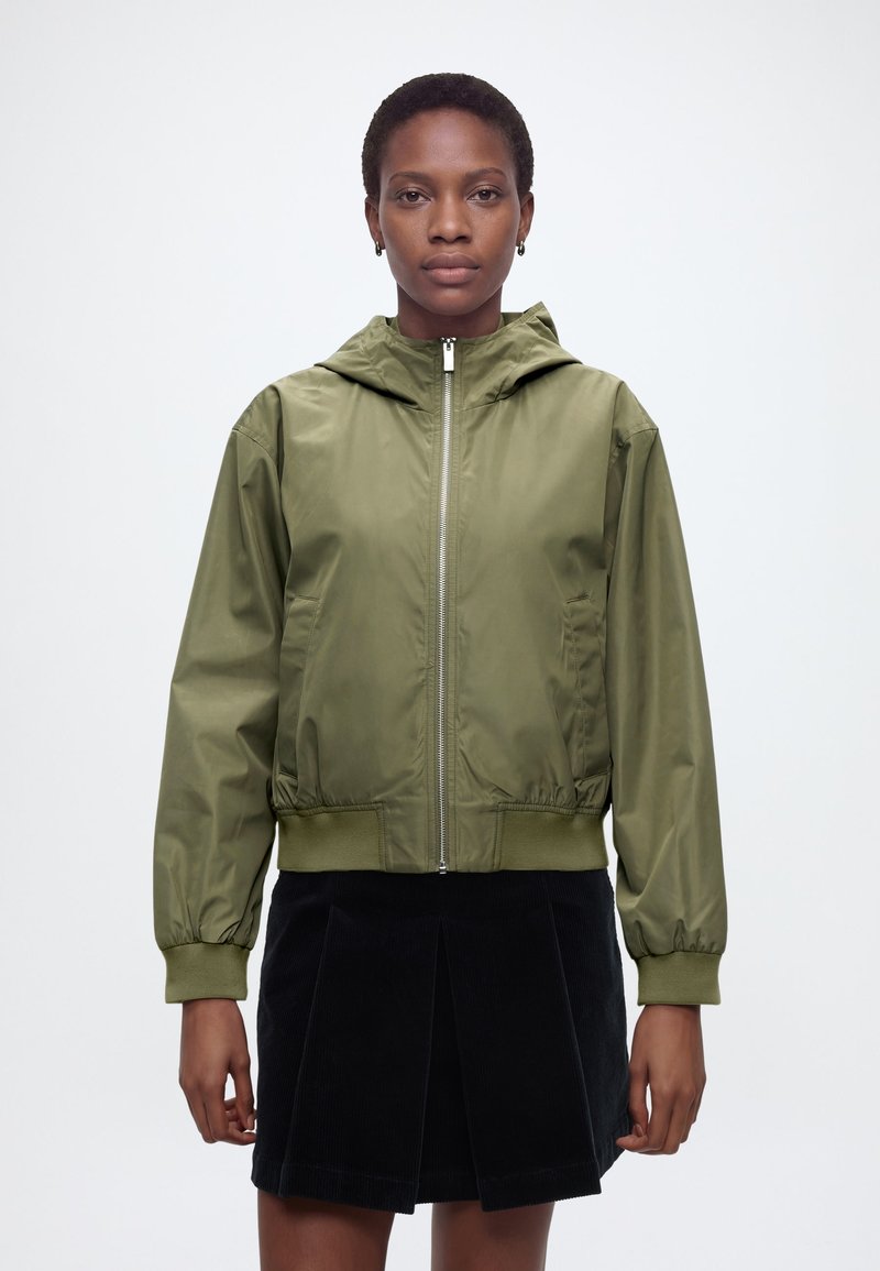 Γυναίκα με πράσινο χακί μπουφάν με φερμουάρ και κουκούλα, και μαύρη πλισέ φούστα, στέκεται μπροστά σε απλό λευκό φόντο.