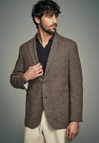 Brauner Hahnentritt-Blazer mit einer Zwei-Knopf-Verschluss, Reverskragen und Fronttaschen, kombiniert mit einem dunklen V-Ausschnitt-Shirt und hellen Hosen.