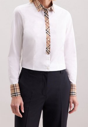 Mujer que lleva una camisa blanca abotonada con cuello, puños y tapeta de botones a cuadros beige, combinada con pantalones negros de talle alto.