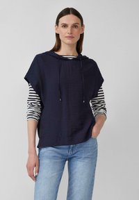Marineblaue, kurzärmlige Kapuzenjacke mit Kordelzügen über einem schwarz-weißen gestreiften Langarmshirt, kombiniert mit hellblauen Jeans.