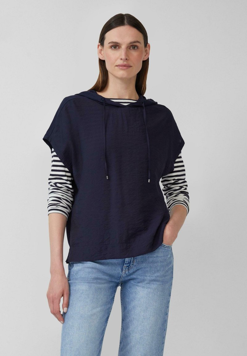 Marineblaue, kurzärmlige Kapuzenjacke mit Kordelzügen über einem schwarz-weißen gestreiften Langarmshirt, kombiniert mit hellblauen Jeans.