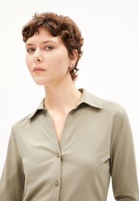Camicia verde oliva con colletto e maniche lunghe, caratterizzata da una patta e dettagli con bottoni. Texture liscia e elementi di design minimi.