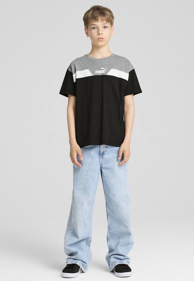 Chemise à manches courtes noire et grise avec une rayure blanche, arborant un logo. Associée à un jean large bleu clair et des baskets noires.