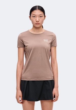 Jeune femme aux cheveux noirs raides porte un t-shirt marron cintré à manches courtes avec logo et une jupe plissée noire, debout sur un fond uni.