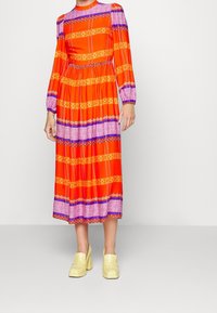 Robe à manches longues rouge et orange avec des motifs violets et jaunes, taille cintrée, jupe fluide. Portée avec des chaussures compensées jaunes épaisses.
