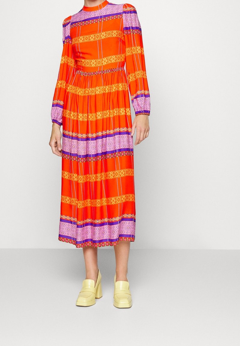 Robe à manches longues rouge et orange avec des motifs violets et jaunes, taille cintrée, jupe fluide. Portée avec des chaussures compensées jaunes épaisses.