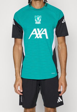 Personne portant un maillot d'entraînement bleu sarcelle et noir du Liverpool Football Club et un short noir Adidas avec des logos visibles sur le maillot et le short.
