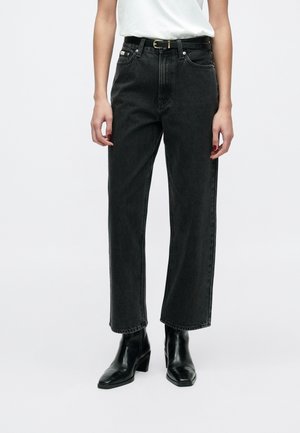 Vrouw draagt zwarte jeans met hoge taille en zwarte riem, wit ingestopt topje en zwarte enkellaarzen met hak, staand tegen een witte achtergrond.