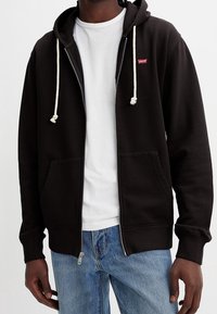 Zwarte sweatshirt met rits en witte koorden, voorzakken en een klein rood logo. Draagt over een wit T-shirt en blauwe jeans.