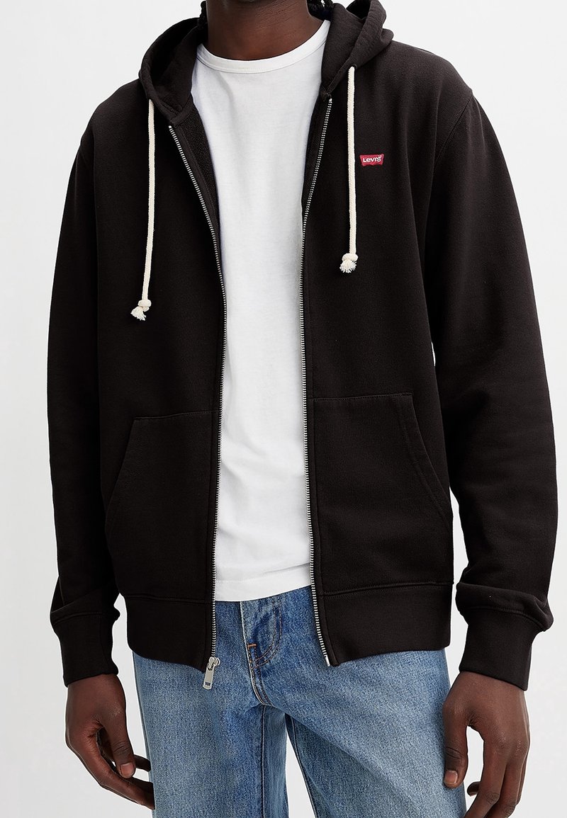 Zwarte sweatshirt met rits en witte koorden, voorzakken en een klein rood logo. Draagt over een wit T-shirt en blauwe jeans.