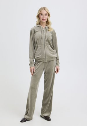 Blondynka stoi ubrana w jasnoszarą welurową bluzę z kapturem na zamek oraz pasujące szerokie spodnie i czarne buty na jednolitym jasnym tle.