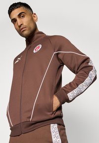 Chaqueta marrón con cremallera, con acentos blancos, que presenta un logo en el pecho y un borde estampado a lo largo de las mangas. Hecha de un tejido suave.