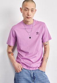 Lila pamut póló rövid ujjal és kerek nyakkivágással, fekete Adidas logóval a mellrészen. Kék farmer sorttal viselve.