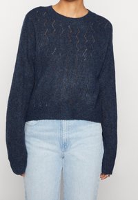 Pull en tricot bleu marine avec un motif zigzag texturé, col rond et manches longues, associé à un jean taille haute bleu clair.