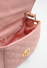 Růžová prošívaná kabelka s hladkým umělým koženým exteriérem, zlatými kovovými prvky a zipovou kapsou uvnitř. Disponuje visící řetízkovou ozdobou.