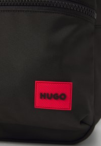 Črna teksturirana tkaninska torba z rdečim pravokotnim patchem, na katerem je v bold črnih črkah napisano "HUGO" blizu zadrge.