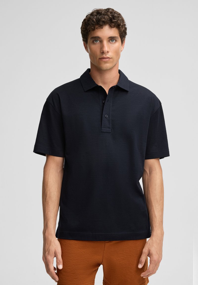 Dunkelblaues Poloshirt mit kragen, kurzen Ärmeln, strukturiertem Stoff und einer Knopfleiste mit drei Knöpfen. Kombiniert mit orangen strukturierten Shorts.