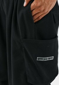 Pantaloni da lavoro neri realizzati in tessuto testurizzato, dotati di una tasca laterale con un logo argentato. Il design presenta sottili dettagli di cucitura obliqua.