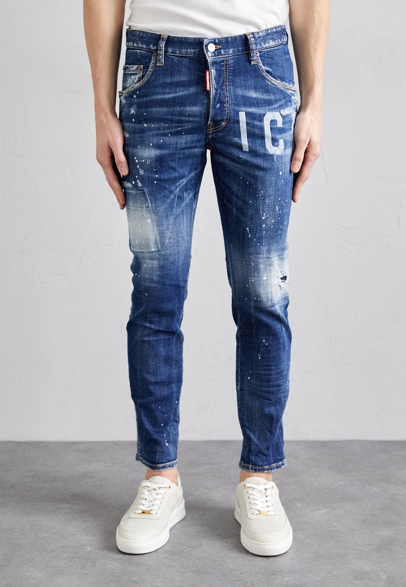 Blaue Denim-Jeans im Slim-Fit mit zerstörten Stellen, Farbspritzmustern und einem weißen Grafikdruck. Kombiniert mit weißen Turnschuhen.