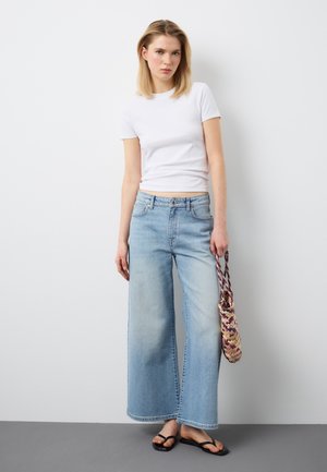 Jonge vrouw draagt een strak wit T-shirt, lichtblauwe jeans met wijde pijpen, zwarte sandalen, houdt een gevlochten tas vast en staat tegen een effen witte muur.