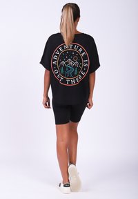 Schwarzes, übergroßes T-Shirt mit einem bunten Grafikdesign von Bergen, Bäumen und dem Text "ADVENTURE IS OUT THERE" auf dem Rücken. Kombiniert mit schwarzen Shorts.