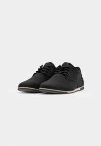 Zapatos negros de cordones, confeccionados en un material suave, con puntera redondeada, diseño minimalista y suela de color claro con una sutil línea de acento.