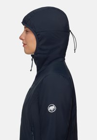 Person som bär en marinblå huva jacka med justerbara huva snören och en vit mammutlogotyp på övre ärmen, vänd mot vänster.