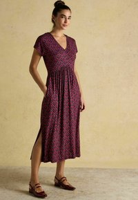 Maxikleid mit V-Ausschnitt, kurzen Ärmeln und gerafftem Bund. Es zeigt ein pinkes Muster auf einem dunklen Hintergrund, mit seitlichen Schlitzen. Braune Absatzschuhe.