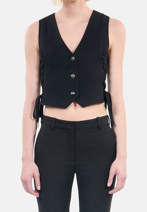 Kvinde iført en sort ærmeløs cropped vest med knapper og sidestropper, kombineret med høj taljede sorte bukser mod en ensfarvet baggrund.