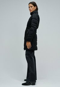 Chaqueta acolchada negra con cierre de cremallera, cuello alto, cintura elástica y bolsillos laterales, combinada con vaqueros oscuros de pierna ancha y botines.