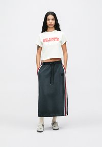 Λευκό κοντομάνικο μπλουζάκι με το "AIR JORDAN FIGHT CLUB" σε κόκκινο χρώμα, συνδυασμένο με μια σκούρα γκρι μακριά φούστα που διαθέτει κόκκινες και ανοιχτό μπλέ ρίγες και τσέπες.