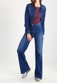 Giacca di jeans con due tasche sul petto, abbinata a una camicia a righe rossa e blu navy. Jeans a zampa d'elefante blu con vita media e scarpe argento.