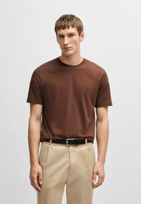 T-shirt marrone a girocollo in tessuto di cotone morbido, con un sottile logo del marchio sul petto. Abbinato a pantaloni chiari e una cintura nera.