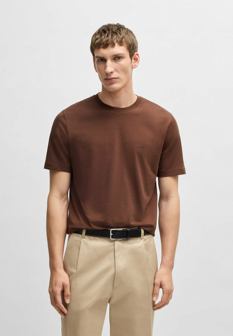 T-shirt marrone a girocollo in tessuto di cotone morbido, con un sottile logo del marchio sul petto. Abbinato a pantaloni chiari e una cintura nera.