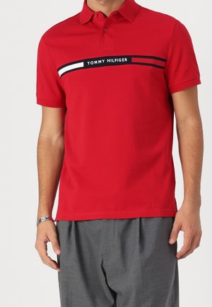 Mand iført en rød Tommy Hilfiger polo med logo og striber over brystet, kombineret med grå bukser og et sølvfarvet armbåndsur.
