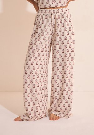 Luźne kremowe spodnie z małymi czerwonymi kwiatowymi wzorami, elastycznym paskiem w talii, noszone boso z rękami opartymi na biodrach.