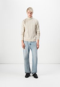 Maglione a collo alto in lavorazione a trecce crema, abbinato a jeans a gamba larga azzurro chiaro e scarpe nere. Lo sfondo semplice mette in risalto i dettagli dell'outfit.