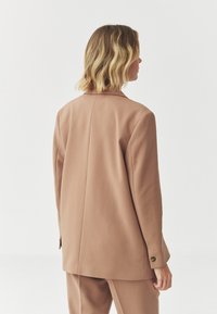 Blazer color cammello con vestibilità sartoriale, spalle strutturate e chiusura a un bottone, realizzato in un tessuto liscio con una leggera texture.