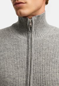 Szary sweter zapinany na zamek błyskawiczny z wysokim kołnierzem, z metalowym zamkiem i teksturowanym wzorem dzianiny, podkreślający miękki i przytulny wygląd.