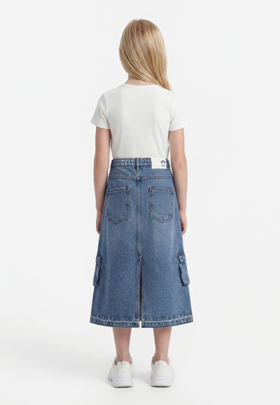 Bērns ar gariem blondiem matiem, valkā baltu īsroku krekliņu un zilu džinsa midi svārku ar kabatām aizmugurē un baltas sporta kurpes, stāv ar muguru pret skatītāju.