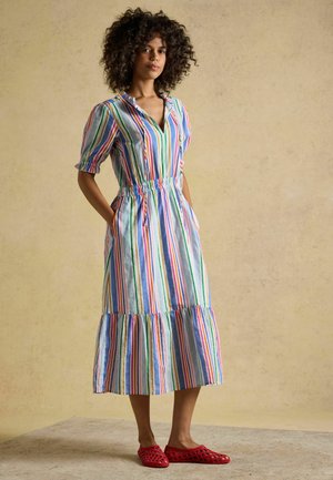 Frau mit lockigem Haar steht mit den Händen in den Taschen, trägt ein bunt gestreiftes Midikleid und rote gewebte Slip-On-Schuhe.