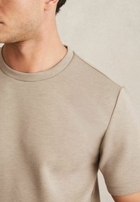 Beige kereknyakú póló, puha anyagból készült. Rövid ujjú, sima textúrával és tiszta varrással. Nincsenek látható minták vagy díszítések.