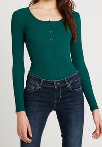 Bodysuit vert côtelé à manches longues avec un décolleté arrondi et une patte de boutonnage à trois boutons, associé à un jean bleu foncé. Texture lisse, design ajusté.