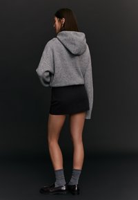 Donna di spalle che indossa un maglione grigio con cappuccio, una minigonna nera, calzini grigi e mocassini marrone scuro su uno sfondo nero.