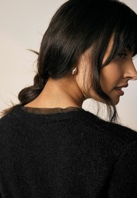 Pull noir texturé avec un col noir transparent, porté avec les cheveux en chignon décontracté, mettant en valeur une simple boucle d'oreille. Gros plan sur le dos.