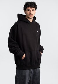 Sudadera negra con capucha, con un gran bolsillo frontal, corte holgado, tejido suave y un pequeño logo blanco en el pecho.